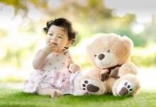 Smiling baby girl sits on grass with a plush teddy bear, enjoying a sunny day.