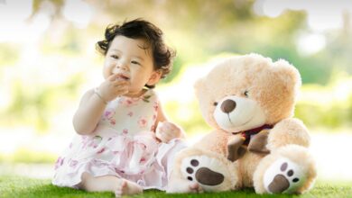 Smiling baby girl sits on grass with a plush teddy bear, enjoying a sunny day.