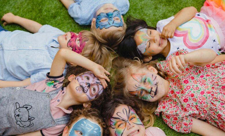 Group of children enjoying an outdoor party with colorful face paint, lying on grass.